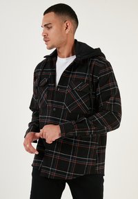 Camicia di flanella a quadri nera e rossa con cappuccio, con chiusura a bottoni frontalmente e due tasche sul petto, indossata da un modello.