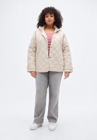 Bež prešita jakna s kapuco, sprednjimi zapenjalnimi jezički in stranskimi žepi, oblečena čez rdeče-belo črtasto majico, kombinirana s sivimi kordurovnimi hlačami.