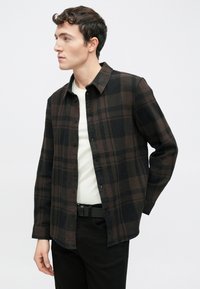 Čierna a hnedá kockovaná flanelová košeľa s zapínaním na gombíky, vybavená špicatým golierom a dlhými rukávmi, nosená cez biele tričko.