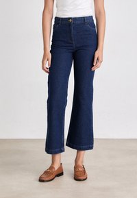 Jeans a vita alta, di colore blu scuro, svasati con tasche anteriori, cuciture e chiusura a bottone, abbinati a mocassini marroni e una camicia bianca aderente.