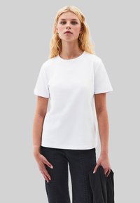 T-shirt en coton blanc à manches courtes et col rond, associé à un jean en denim noir. Le mannequin tient un vêtement noir dans une main.