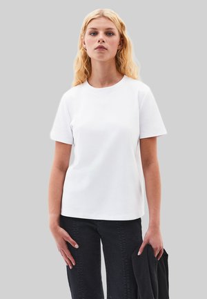T-shirt en coton blanc à manches courtes et col rond, associé à un jean en denim noir. Le mannequin tient un vêtement noir dans une main.
