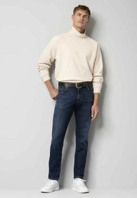 Crème sweatshirt met hoge kraag, donkerblauwe jeans, zwarte riem en witte sneakers. Glad materiaal en aangesloten ontwerp. Staande pose tegen een lichte achtergrond.