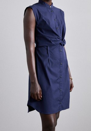 Mouwloze marineblauwe jurk met knopen en een knoopdetail in de taille, gedragen door een persoon met een donkere huid tegen een effen achtergrond.