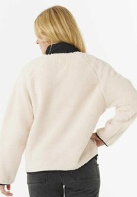 Vrouw met blond haar die een crèmekleurige fleecejas draagt met een zwarte kraag en manchetten, staande met één hand op de heup, met de rug naar de kijker gekeerd.