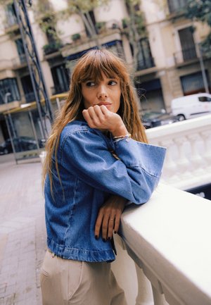 Giacca di denim con taglio morbido e maniche larghe, indossata sopra pantaloni chiari. Al polso si notano braccialetti distintivi, con uno sfondo testurizzato.