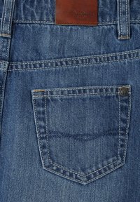 Temno modra denim tkanina z majhnim zadnjim žepom, svetlo rjava usnjena etiketa prišita zgoraj, ter kontrastno bela šivanka ob šivih.