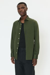 Donkergroen shirt met lange mouwen, voorzien van een knoopsluiting aan de voorkant, puntige kraag en een duidelijke stofstructuur. Draagt over een zwart shirt en gecombineerd met donkere broek.