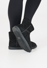 Botas de ante negras hasta el tobillo con una suela de goma texturizada, presentan un diseño cosido y un corte bajo, mostradas desde atrás y de lado.