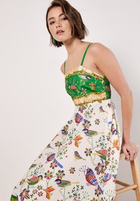 Abito multicolore con un corpetto verde a fantasia e una gonna fluida color crema con illustrazioni floreali e di uccelli. Spalline a spaghetti e vita aderente.