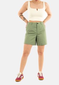 Shorts in cotone verde con taglio sartoriale, dotati di passanti per la cintura e tasche, abbinati a un top corto bianco a costine e scarpe da ginnastica bordeaux.