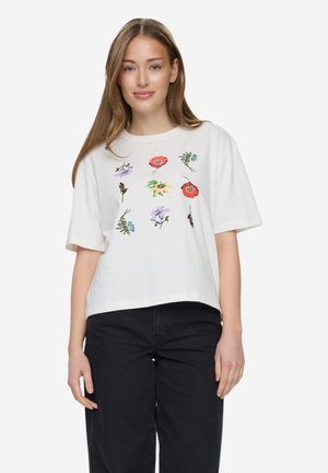 Młoda kobieta w białej koszulce z kolorowymi haftowanymi kwiatami i czarnych spodniach, stojąca na białym tle.