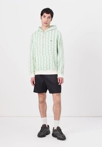 Felpa verde chiaro con un motivo geometrico, polsini crema, pantaloni corti neri e scarpe da corsa scure. Il modello ha i capelli corti e un tatuaggio sulla gamba.