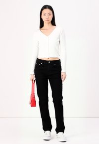 Λευκή ριγέ κ cropped μπλούζα με μακριά μανίκια και κουμπιά, συνδυασμένη με μαύρα τζιν ευθείας γραμμής και λευκά αθλητικά παπούτσια. Κρατάει μια ροζ τσάντα ώμου.