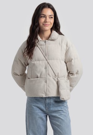 Junge Frau mit langen dunklen Haaren, die eine beige Daunenjacke und hellblaue Jeans trägt, steht mit den Händen hinter dem Rücken.