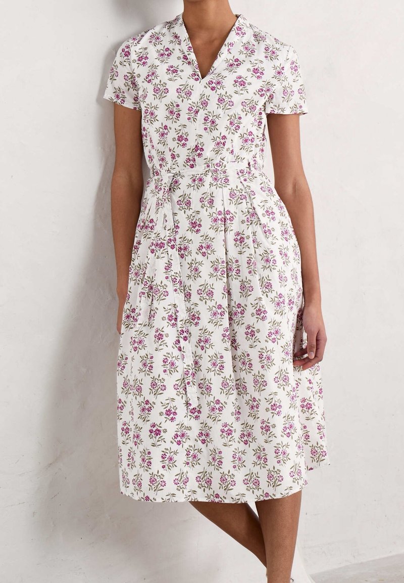 Femme portant une robe midi blanche avec un motif floral rose, manches courtes, col en V et ceinture assortie en tissu nouée à la taille.