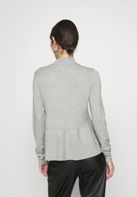 Kobieta z ciemnymi włosami związanymi w niski kok, nosząca srebrne kolczyki w kształcie kół, jasnoszary sweter z długim rękawem i rozkloszowanym dołem oraz czarne spodnie, zwrócona plecami.