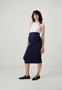 Raska kvinna med axellångt mörkt hår klädd i en ärmlös vit topp, marinblå figursyt skirt och svarta platta skor.