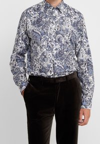 Mężczyzna ubrany w niebieską i białą kwiecistą koszulę w paisley z długim rękawem, włożoną w ciemnobrązowe aksamitne spodnie, z jedną ręką w kieszeni.