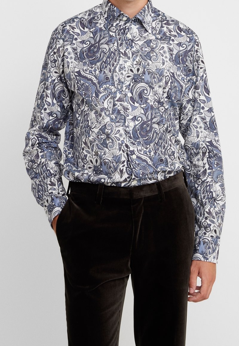 Mężczyzna ubrany w niebieską i białą kwiecistą koszulę w paisley z długim rękawem, włożoną w ciemnobrązowe aksamitne spodnie, z jedną ręką w kieszeni.