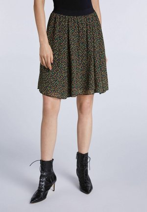 Femme portant une jupe noire à fleurs longueur genou associée à des bottines à talons noires à lacets, sur un fond uni.