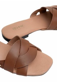 Brązowe skórzane sandały z płaską podeszwą, z krzyżującymi się paskami i teksturowaną wkładką. Czarna podeszwa zewnętrzna zapewniająca przyczepność i stabilność.