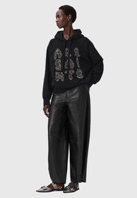 Czarna bluza z kapturem z dużym srebrnym tekstem graficznym, zestawiona z szerokimi czarnymi skórzanymi spodniami oraz metalicznymi płaskimi butami. Proste tło.