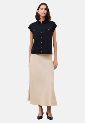 Femme portant une chemise noire à manches courtes avec des poches et une jupe beige longueur cheville, debout en talons noirs.