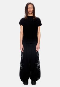 Άτομο με dreadlocks στέκεται φορώντας μαύρο φθαρμένο t-shirt και μαύρο φαρδύ παντελόνι με σχέδια, μαζί με μαύρα αθλητικά παπούτσια, σε λευκό φόντο.