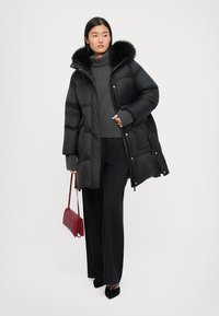 Black puffer jacket with a detachable fur collar, zip and snap closures, worn over a dark gray sweater and black wide-leg trousers. Red handbag.