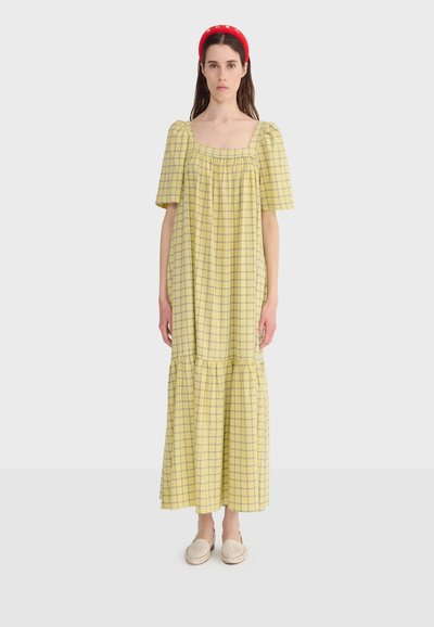 Femme portant une robe midi ample à carreaux jaunes avec des manches bouffantes, un bandeau rouge et des chaussures beige à enfiler, debout devant un fond blanc.