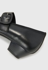 Black leather shoe with a curved design, subtle sheen, and a square heel. Features a buckle strap and detailed stitching along the edge.