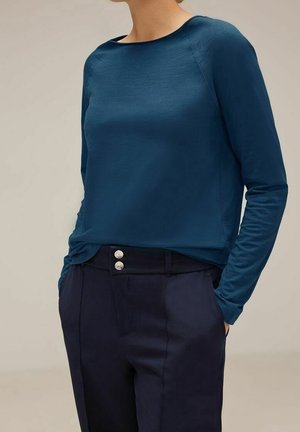 Person trägt ein langärmeliges marineblaues Hemd, das in eine hochgeschnittene dunkelblaue Hose mit silbernen Doppelknöpfen gesteckt ist, die Hände in den Taschen.