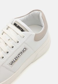 White leather sneaker with a beige accent at the heel, featuring a textured surface, round laces, and black logo detail on the side.