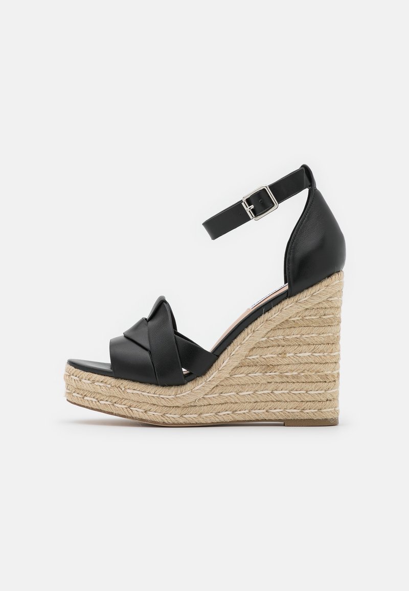 Sandale compensée en cuir noir avec plateforme en jute tressé, brides croisées à l'avant et fermeture à boucle à la cheville, vue de profil sur fond blanc.