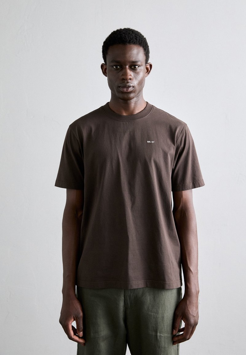 T-shirt en coton marron avec un col rond, des manches courtes, et un petit logo blanc sur la poitrine, associé à un pantalon vert.