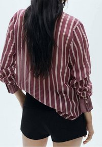 Blouse rayée bordeaux à manches longues, présentant des lignes verticales blanches. Le tissu semble léger, associé à un short noir.