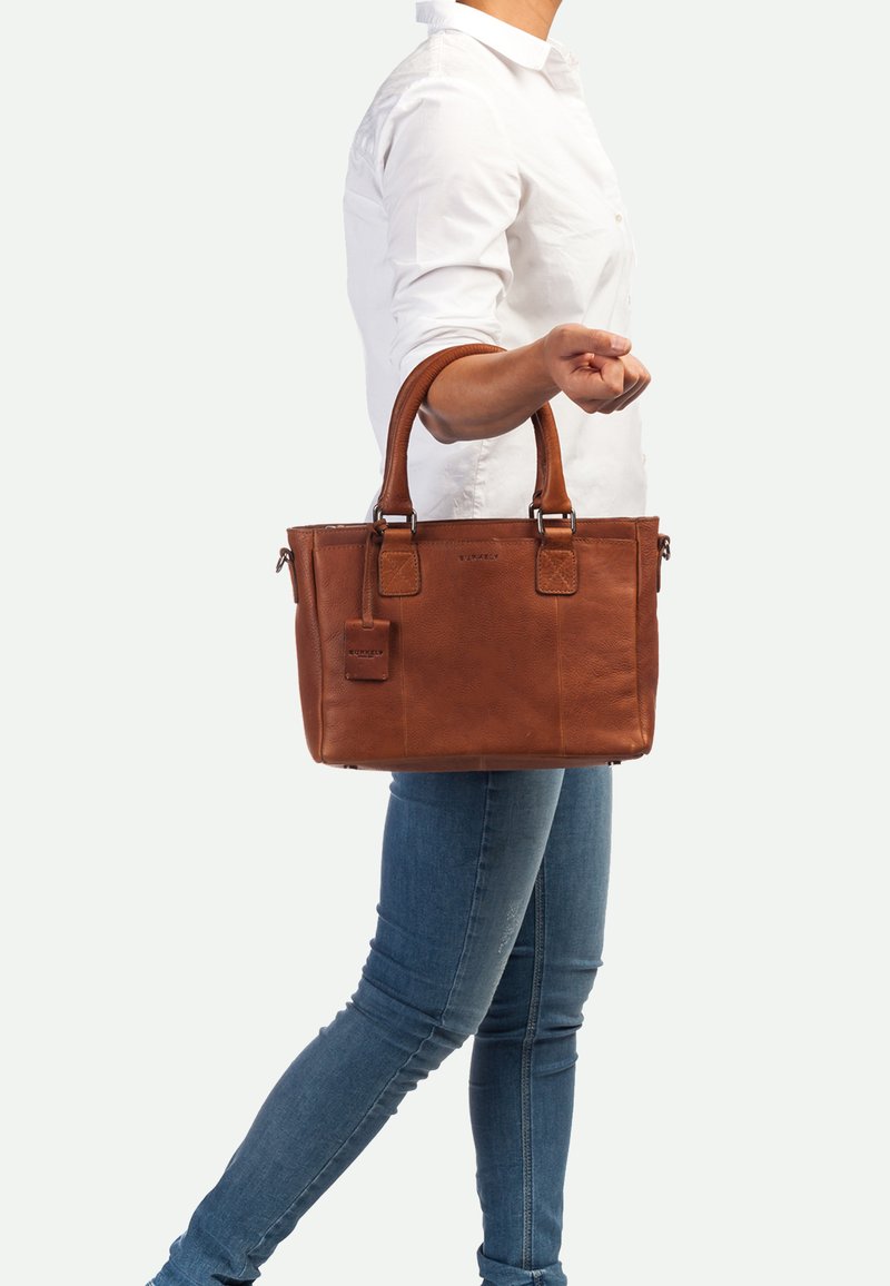 Sac à main en cuir marron avec deux poignées supérieures, forme rectangulaire, finition texturée et étiquette de bagage attachée. Accents brodés sur les bords.