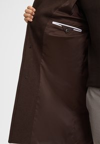 Bruine wollen jas met een gladde, chocoladekleurige voering. Heeft een knoopsluiting en een binnenzak met witte accentstiksel.