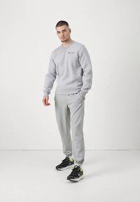 Sivá mikina s malým logom, skombinovaná s svetlosivými teplákmi. Čierne tenisky s neónovými akcentmi. Interiér s jednoduchým pozadím.