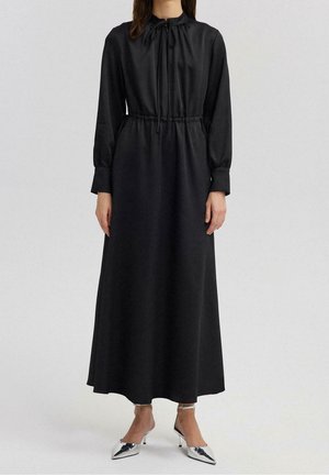 Vrouw draagt een lange zwarte jurk met een koordsluiting bij de halslijn en taille, lange mouwen, gecombineerd met zilverkleurige schoenen met spits toelopende neus.