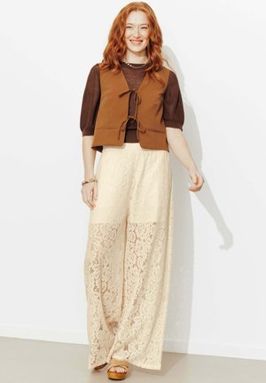 Femme rousse souriante portant un pull marron à manches courtes, un gilet noué devant couleur rouille, un pantalon large en dentelle crème et des sandales, debout contre un mur blanc.