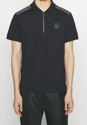 Mand iført sort poloshirt med korte ærmer og lynlås foran, grå skulderpaneler og lille brystlogo, kombineret med sorte bukser.