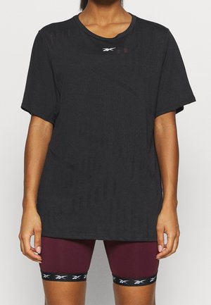 Personne portant un t-shirt Reebok noir ample et un cuissard de cyclisme moulant bordeaux avec des bandes noires du logo Reebok à la cuisse.
