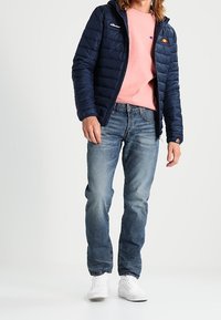 Giacca puffer blu marina con cappuccio, maglione rosa, jeans blu e sneakers bianche. La giacca presenta un logo sul petto e una texture trapuntata.