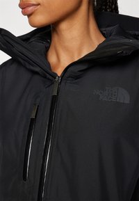 Crna vodootporna jakna s visokim ovratnikom, dvojim patentnim zatvaračima i logom North Face u mat printu. Glatka tekstura, elegantan dizajn.