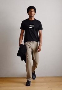 Black cotton T-shirt with a white Coca-Cola logo, beige trousers, and black shoes. The figure holds a black jacket while walking.