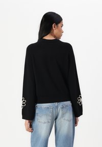 Svart genser med lange ermer som har en blomsterbrodert design på mansjettene. Kombinert med lyseblå, avslappede jeans.