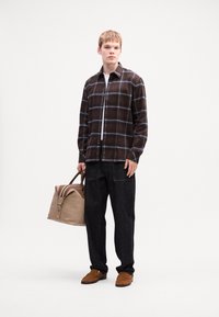 Junger Mann in braunem karierter Hemd und schwarzen Hosen, der eine beige und braune Leder Tasche hält, stehend vor weißem Hintergrund.