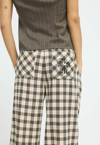 Weite Karottenhosen im Karomuster in Beige, Schwarz und Braun. Mit zwei großen aufgesetzten Taschen mit bestickten Details und aus strukturiertem Stoff.